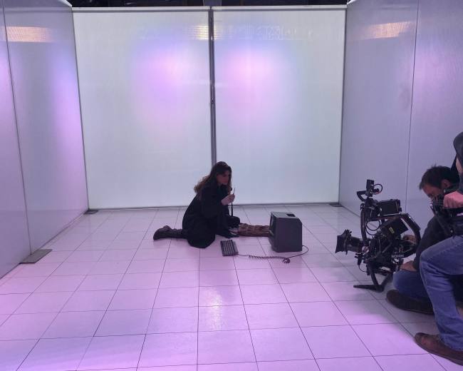 A woman sits on the floor in a sparse, white room, working on a small computer while a crew member adjusts camera equipment nearby.