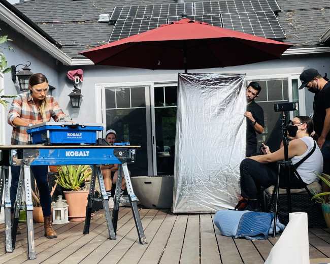 A woman works on a project at a table in a backyard while several people observe and assist.