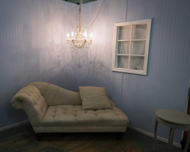 The image shows a lightly colored chaise lounge with a throw pillow, positioned near a window and a chandelier on the wall in a softly lit corner of a room.