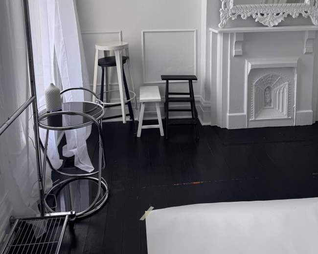 The image shows a bright room with white walls, a large ornate mirror, three stools in different colors, a clothing rack, and a round table.