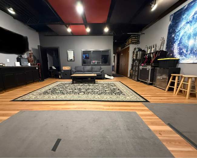 The image shows a studio or rehearsal space with a wooden floor, a large area rug, a couch, and music equipment in the background.