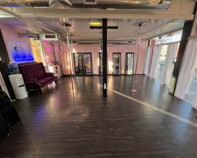 The image shows an interior space designed for pole dancing, featuring mirrored poles, a seating area with a pink couch, and large windows providing natural light.
