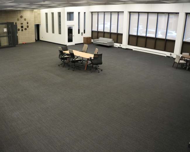 An empty, well-lit room features several tables and chairs arranged in the center, with large windows along one wall and an elevator to one side.