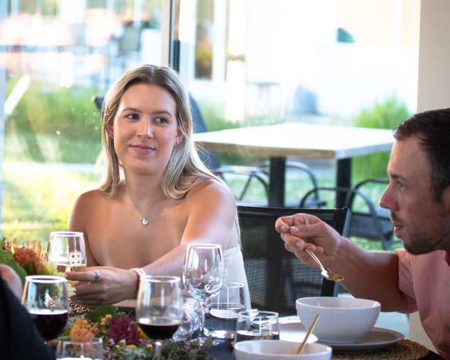 A woman and a man are seated at a dining table with glassware and bowls, engaged in conversation in a bright restaurant setting.