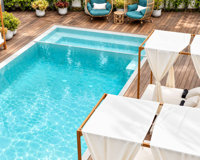 The image shows a modern outdoor space featuring a swimming pool, lounge chairs with canopies, and seating arrangements on a wooden deck surrounded by greenery.