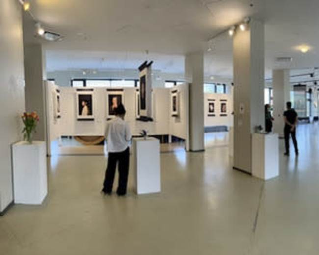The image shows an art gallery with several framed portraits displayed on white walls, while visitors explore the space amidst a few pedestals and seating arrangements.