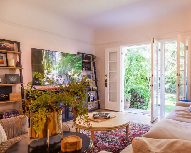 A cozy living room features a large television, a round coffee table, and a door open to a lush garden.