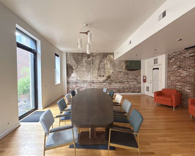 A long conference table with chairs is set up in a room featuring exposed brick walls and a large window leading to an outdoor space.