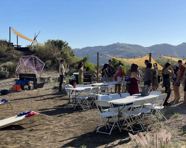 A group of people gathers around tables set up outdoors on a hillside with a view of mountains in the background.