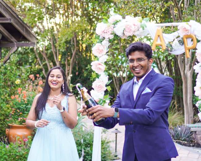 A man in a suit opens a champagne bottle while a woman in a light blue dress smiles beside him, surrounded by floral decorations in a garden setting.