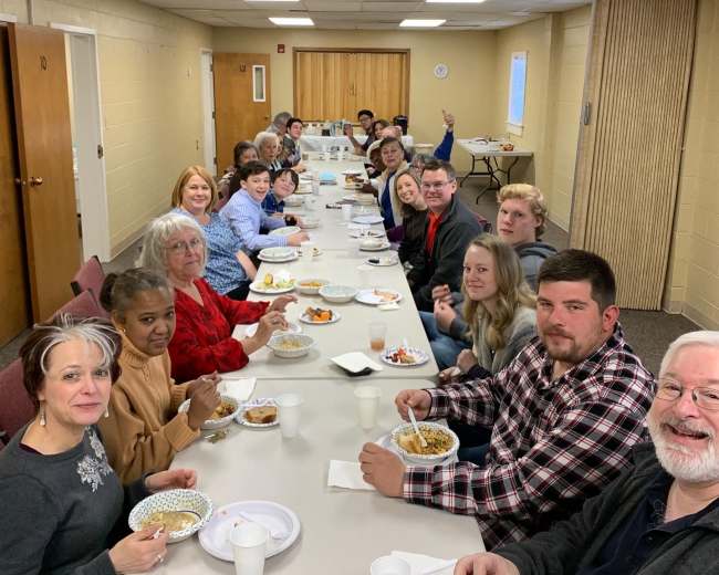 A group of people gathers around a long table, enjoying a meal together in a well-lit room.