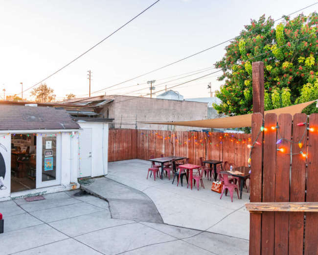 The image shows an outdoor seating area at a record store, featuring tables and chairs surrounded by a wooden fence and a tree, with string lights illuminating the space.