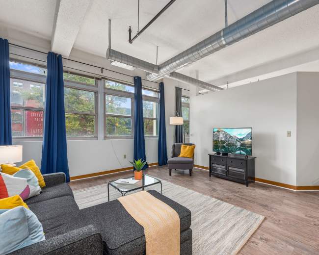 A modern living room features a gray sofa with colorful cushions, a coffee table, a television on a black stand, and large windows with blue curtains.