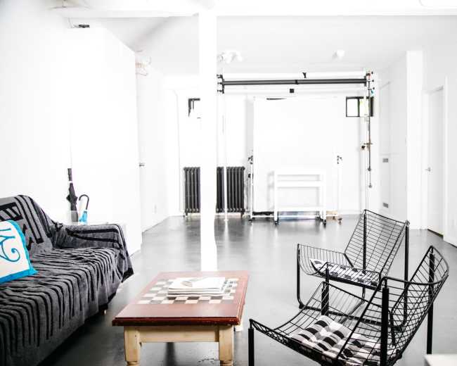 The image shows a minimalist, modern interior featuring a black and white striped sofa, two wireframe chairs, a wooden coffee table with a checkerboard, and a backdrop setup in a bright, white room.