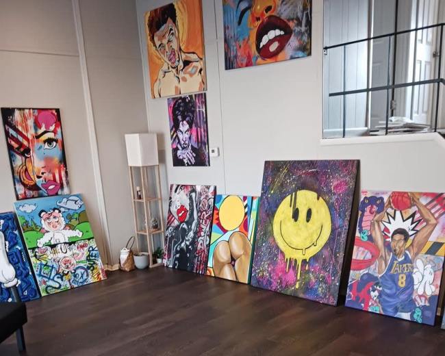 An art studio with several colorful paintings displayed on the walls and propped against the floor, along with a modern lamp and a black chair.