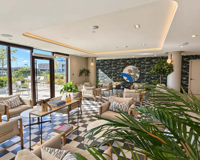 The image shows a modern lounge area with seating arranged around coffee tables, adorned with plants and large windows that allow natural light to fill the space.