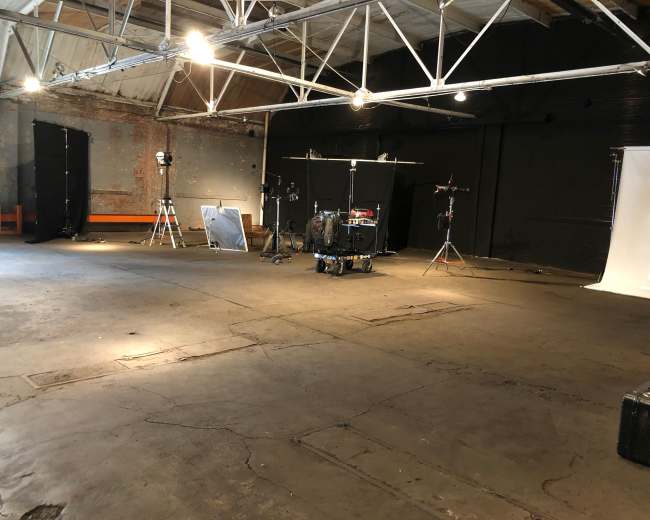 An empty industrial space with a concrete floor, featuring lighting equipment, a drum set, and black backdrops.