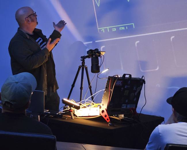 A presenter gestures towards a screen displaying a waveform while speaking to an audience seated in a dimly lit room.