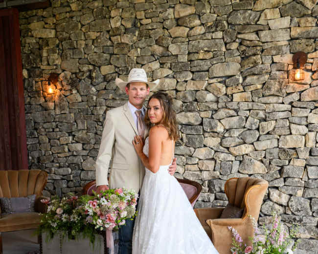 A couple poses together in front of a stone wall, with the woman in a wedding dress and the man in a light suit and hat, surrounded by floral arrangements and vintage furniture.
