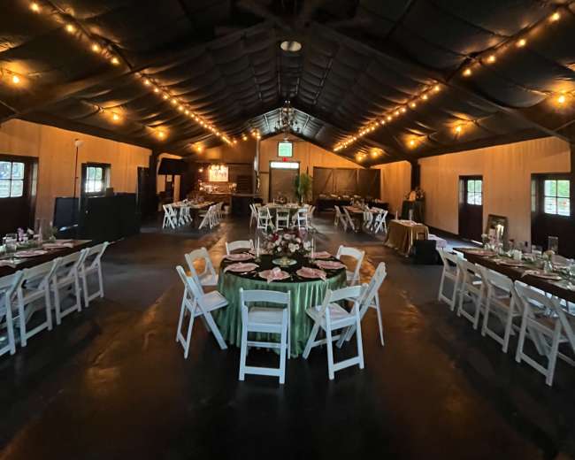The image shows a spacious indoor venue with tables set for a gathering, illuminated by warm string lights along the ceiling.