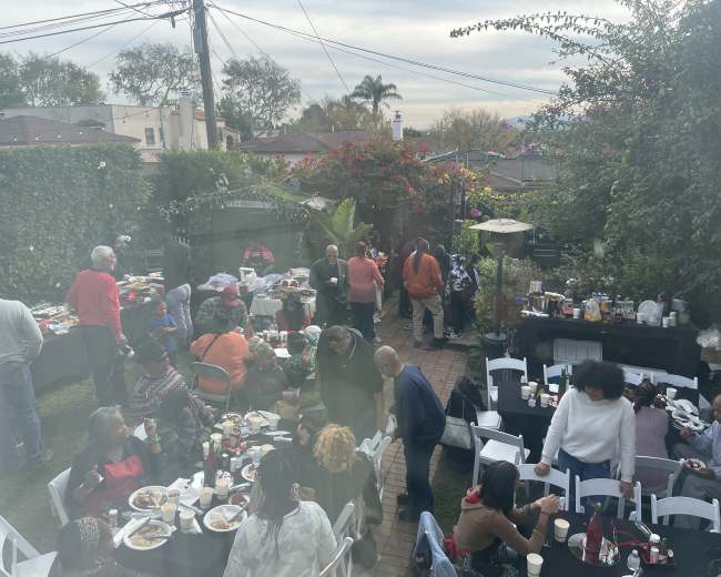 A large group of people gathers in a backyard filled with tables of food, mingling and enjoying a meal outdoors.