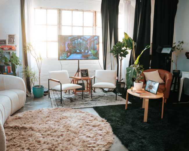 A cozy living room features two armchairs and a sofa, surrounded by several potted plants, with a television displaying a scenic image and natural light streaming through large windows.