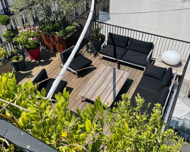The image shows a rooftop terrace with wooden flooring, several black outdoor couches, a coffee table, and potted plants.