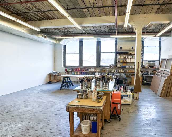 The image shows a spacious art studio with wooden floors, large windows, and various art supplies organized on tables.