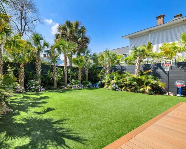 The image shows a landscaped backyard featuring lush greenery, palm trees, and a wooden deck.