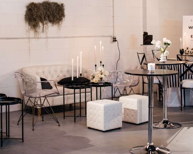 The image shows a modern indoor space featuring a white sofa, tall candle holders with candles, and various styled tables and chairs against a white brick wall.