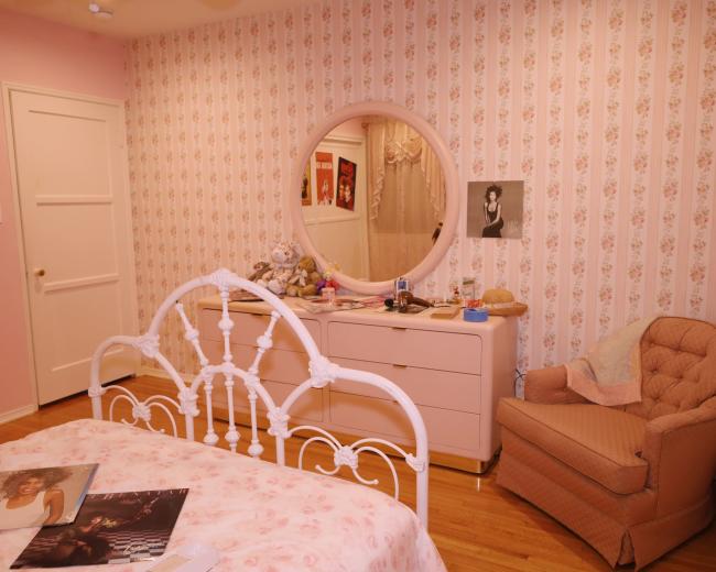 The image shows a bedroom with floral wallpaper, a round mirror above a dresser, and a bed with a pink floral bedspread, accompanied by a chair in the corner.