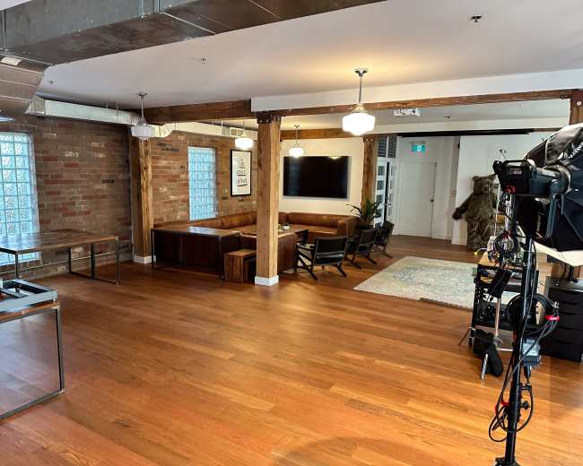 The image shows a spacious modern loft with hardwood floors, exposed brick walls, and a seating area that includes a couch and chairs, alongside a large television and a decorative rug.