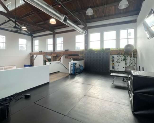 An open gym space with high wooden ceilings, large windows, and various fitness equipment arranged along the walls.