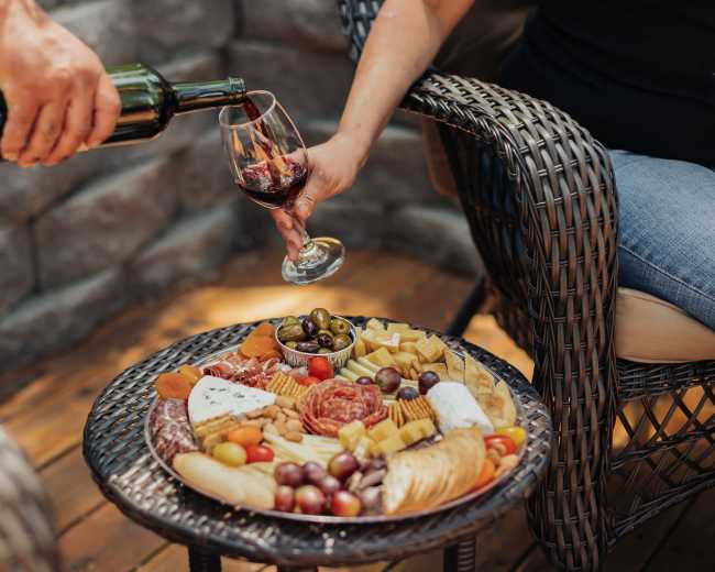 A hand pours red wine into a glass above a table filled with a variety of cheeses, cured meats, olives, and dried fruits.