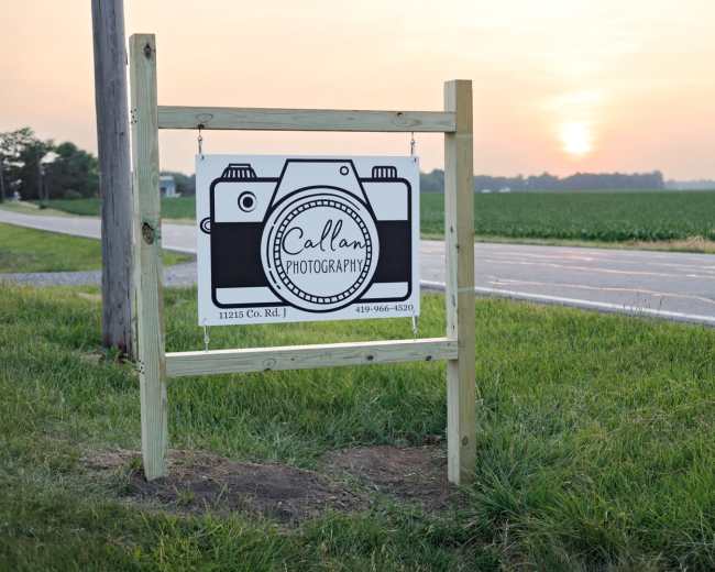 A wooden sign for Callan Photography is posted along a rural road, with a small camera graphic and a sunset in the background.