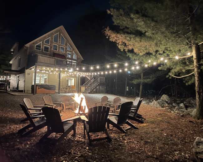 A fire pit surrounded by wooden chairs is set in front of a house illuminated by string lights at night, with trees in the background.