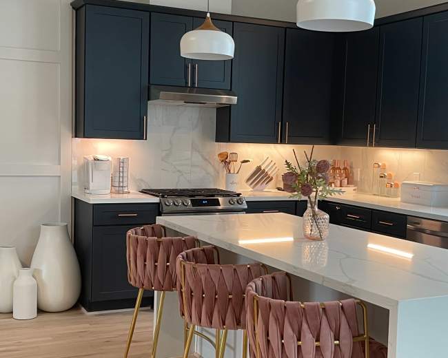 A modern kitchen with dark cabinetry, white countertops, and three pink and gold barstools at a central island.