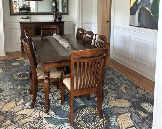 The image shows a dining room featuring a wooden table surrounded by eight chairs, a decorative light fixture overhead, and a large piece of art on the wall.