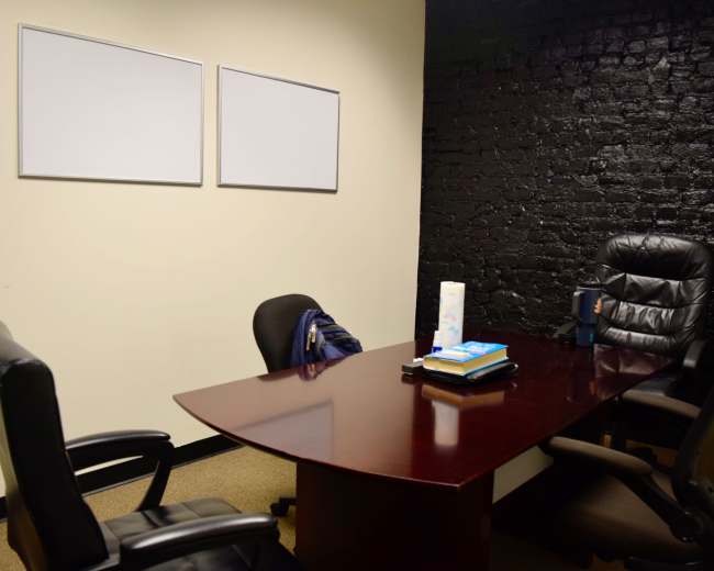 The image shows a small office with a dark brick wall, a wooden table, two office chairs, and two blank whiteboards mounted on the wall.