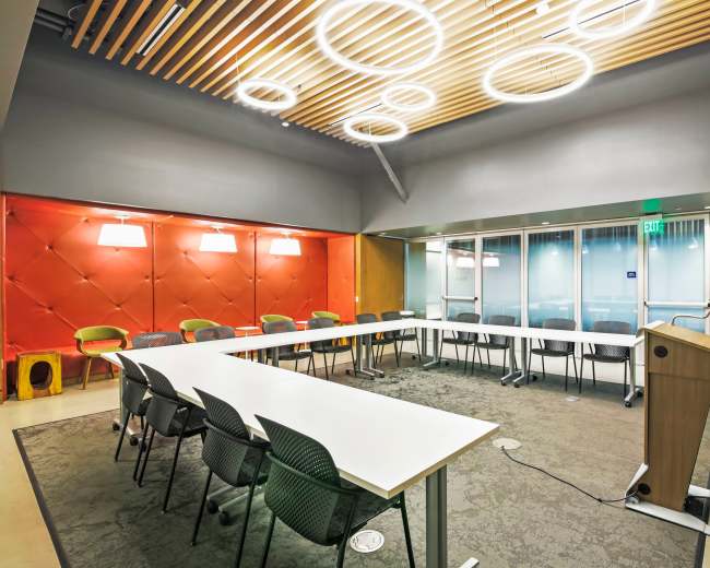 A modern meeting room features a large white table surrounded by gray and green chairs, with a podium and a ceiling of circular light fixtures.