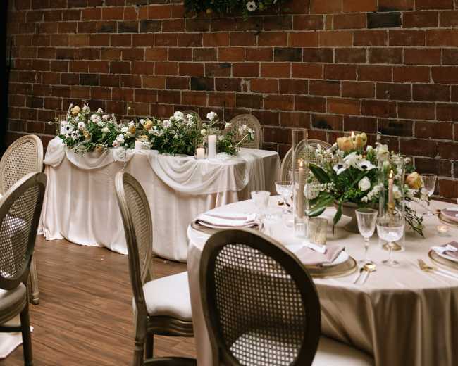 A dining setup with elegantly arranged tables adorned with floral centerpieces and candles, set against a brick wall.