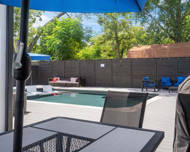 The image depicts a backyard pool area with a swimming pool surrounded by lounge chairs and an outdoor dining table, all enclosed by a wooden fence and shaded by large umbrellas.