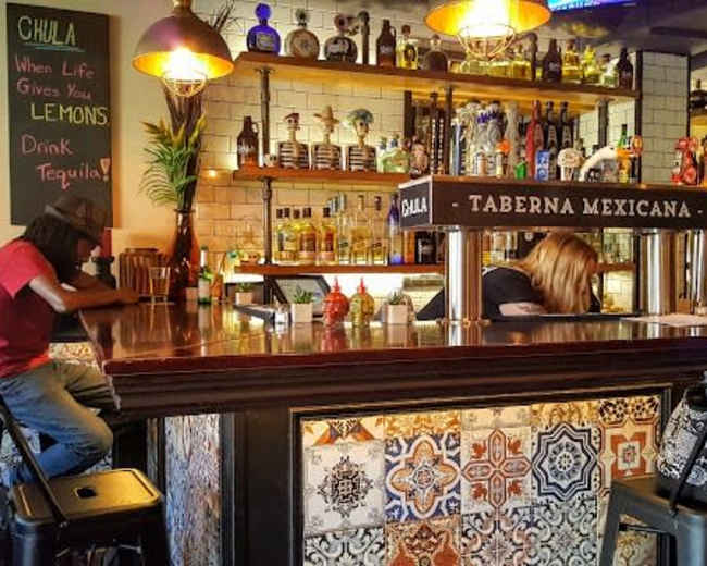 A man sits at the bar while two bartenders prepare drinks in a colorful Mexican restaurant.