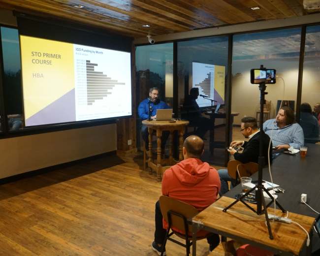 A presenter stands at a table in a modern conference room, displaying a presentation on a screen while three attendees sit in front, engaged in the session.