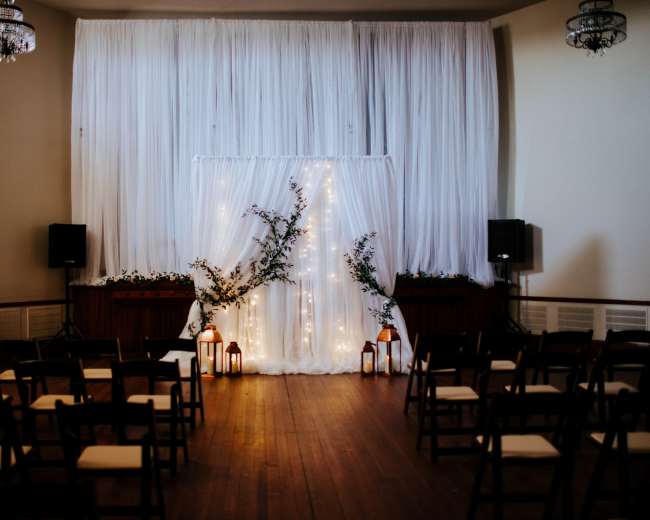 A decorated ceremony space features a white backdrop adorned with fairy lights, flanked by plants, and rows of wooden chairs facing the setup.