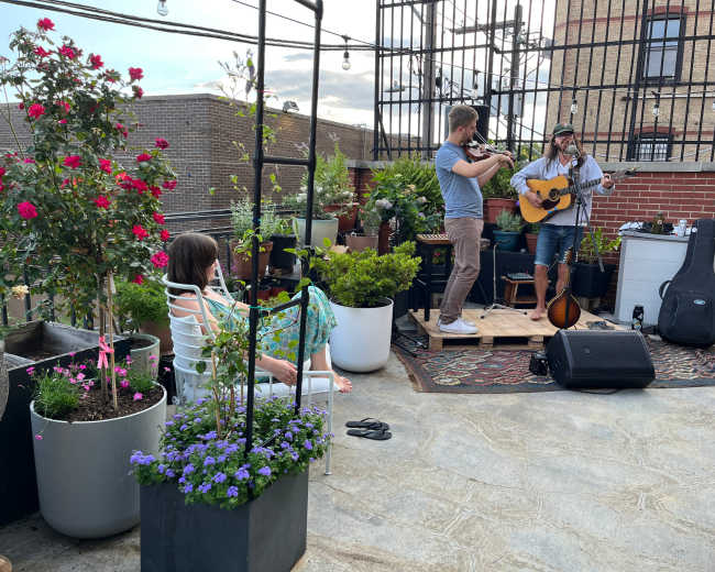 Two musicians perform on a rooftop surrounded by plants, while a woman relaxes in a chair nearby.