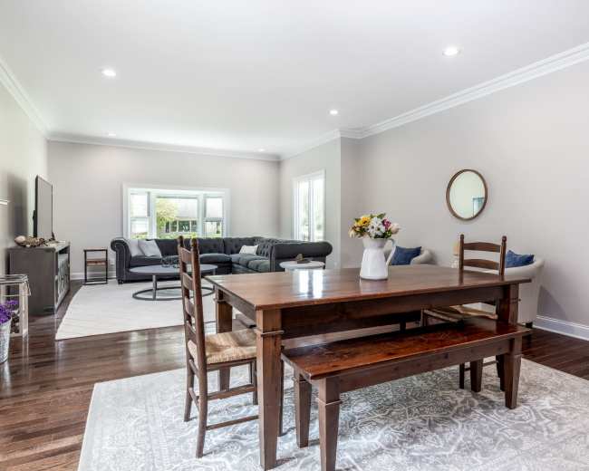 A wooden dining table with chairs sits on a patterned rug in a bright, spacious living area featuring a gray sofa and large windows.