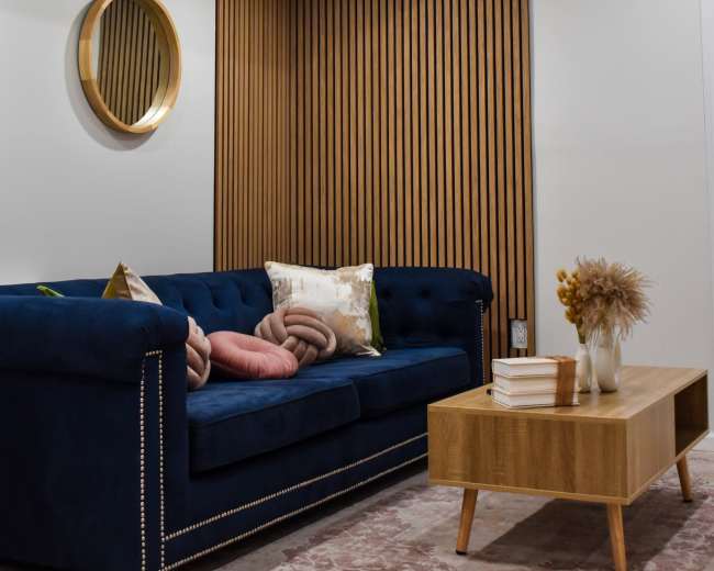 A modern living room features a blue velvet sofa with decorative pillows, a wooden coffee table, and a round mirror above a wooden accent wall.