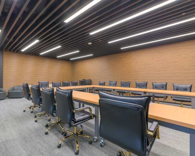 A large, modern conference room features a rectangular table surrounded by ergonomic chairs, with wood-paneled walls and recessed lighting.