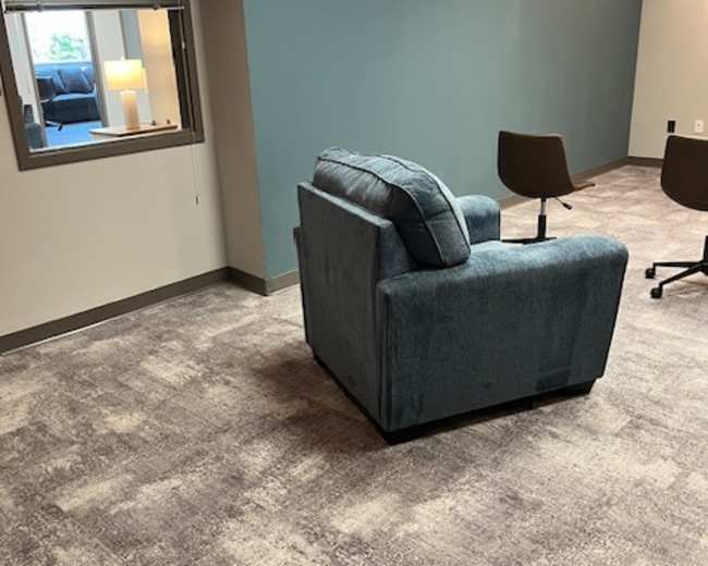 A blue armchair sits in a corner of an office space, next to a window looking out to another room with a lamp.